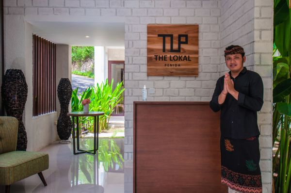 Reception at The Lokale Residences in Bali: smiling Balinese staff in traditional attire greets visitors behind wooden desk amid tropical plants and open courtyard. - Bali Villas